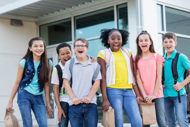 A multi-ethnic group of six middle school students, 11 to 13 years old, standing together outside the school building, carrying backpacks and lunch bags, smiling and laughing at the camera. The boy in the middle wearing eyeglasses has down syndrome.